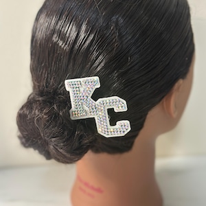 May include: A white hair clip in the shape of the letters "KC" is decorated with iridescent rhinestones. The hair accessory is placed in dark brown hair styled in a low bun. The clip adds a touch of sparkle to the hairstyle.