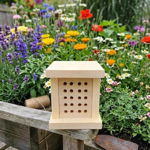May include: A light-colored wooden bee house featuring a square roof and base with a central block containing a grid of twenty circular nesting holes. The structure is constructed from smooth, unfinished light wood with a clean, geometric design.