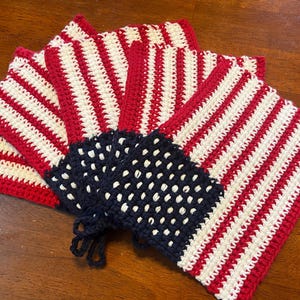 May include: Handmade crochet potholders in a patriotic design. The potholders feature red and white stripes with a blue square containing white dots, resembling the American flag. The set is made of yarn and is suitable for kitchen use.