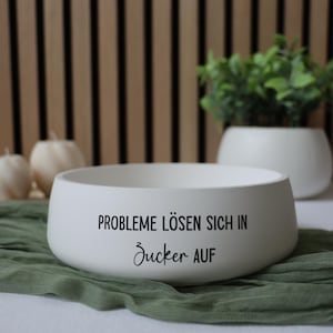 Könnte beinhalten: Eine weiße Keramikschale mit dem deutschen Text "PROBLEME LÖSEN SICH IN ZUCKER AUF" in schwarzer Schrift. Die Schale steht auf einem grünen Tuch, mit einem hölzernen Lattenhintergrund und einem weißen Pflanzgefäß mit Grün.