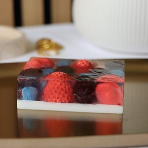 Könnte beinhalten: Ein quadratisches Seifenstück mit eingebetteten Erdbeeren, Blaubeeren und Brombeeren. Die Seife hat ein klares, geleeartiges Aussehen mit einer weißen Basis und liegt auf einem goldenen Tablett. Die Seife ist ein dekorativer Artikel.