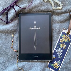 May include: A black and white illustration of a sword on a Kindle e-reader. The e-reader is on a gray fuzzy surface with a floral bookmark and a gold chain with colorful beads.