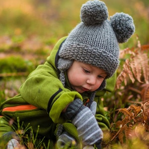 Puede incluir: Un niño pequeño con un gorro de punto gris con pompones y un mono verde está explorando el suelo del bosque. El niño está mirando hacia el suelo y el gorro está tirado sobre su frente.