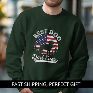 Op de afbeelding: Donkergroene sweatshirt met de witte tekst "BEST DOG Dad Ever". Het ontwerp toont een silhouet van een hond voor een Amerikaanse vlag. De sweatshirt heeft een ronde hals.