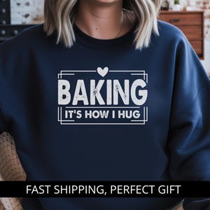 Op de afbeelding: Marineblauwe sweatshirt met de witte tekst "BAKING IT'S HOW I HUG" in een witte rechthoekige lijst, bekroond met een hart. De sweatshirt heeft een effen marineblauwe kleur.