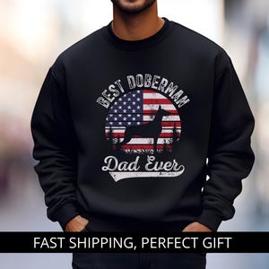 Op de afbeelding: Zwart sweatshirt met een afbeelding van een Dobermann-silhouet, de Amerikaanse vlag en de tekst "Best Doberman Dad Ever". Het ontwerp is in een distressed stijl. Het woord "BLACK" staat in een roze cirkel.