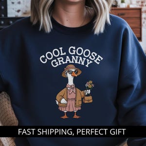 以下が含まれることがあります： 「COOL GOOSE GRANNY」の文字がプリントされたネイビーブルーのスウェットシャツ。サングラス、帽子、ジャケットを身につけ、花とハンドバッグを持った漫画のガチョウが描かれています。カジュアルなスタイルです。
