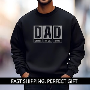 Op de afbeelding: Zwart sweatshirt met de tekst "DAD" in een rechthoekig ontwerp, met de namen "EMMA JACK TOM" eronder. Het sweatshirt is effen zwart. De tekst "FAST SHIPPING, PERFECT GIFT" staat onderaan.