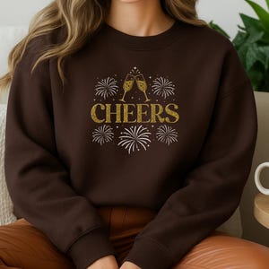 May include: A dark chocolate-colored sweatshirt with the word "CHEERS" in gold glitter lettering. The design includes two champagne glasses clinking and fireworks. A white mug sits on a wooden table.