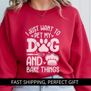 Op de afbeelding: Rode sweatshirt met de witte tekst "I JUST WANT TO PET MY DOG AND BAKE THINGS". Het ontwerp bevat een hondenpootafdruk, een garde en een koksmuts. Een roze cirkel met "RED" staat linksboven.