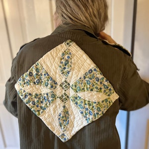 May include: A green utility jacket with a white and blue quilted patch sewn on the back. The patch features a floral design with blue and green flowers.