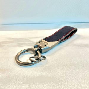 May include: A silver key chain with a black leather strap and red stitching. The strap is attached to a silver ring with a smaller ring attached to it.