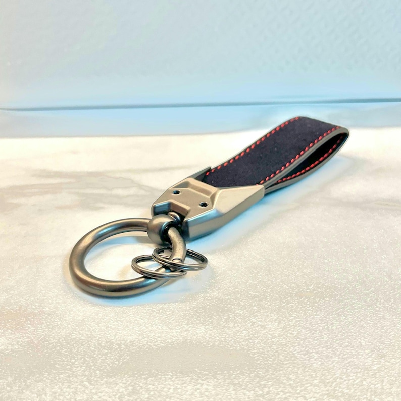 May include: A silver key chain with a black leather strap and red stitching. The strap is attached to a silver ring with a smaller ring attached to it.