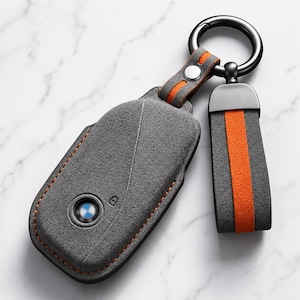 Könnte beinhalten: Ein graues Schlüssel-Etui aus Wildleder mit kreisförmiger Aussparung für das BMW-Logo, orangefarbenen Nähten und einem orange gestreiften Wildlederband an einem schwarzen Metallring. Ein kleines Schloss-Symbol ist auf der grauen Oberfläche.