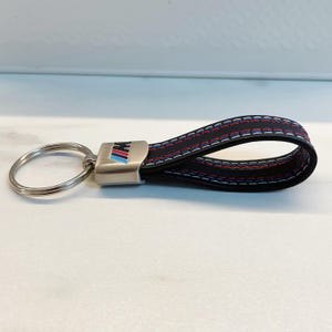 Pode incluir: Um chaveiro de couro preto com um anel de metal prateado e um logotipo BMW M azul, vermelho e branco. A tira de couro tem costuras brancas.
