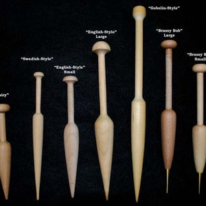 May include: A collection of 10 wooden knitting bobbins in various sizes and styles, including "Gobelin-Style", "English-Style", "Brassy Bob", "Swedish-Style", "Aubusson-Style Bone", "Petite", and "Fairy". The bobbins are arranged in order of size, with the smallest bobbin at the top and the largest bobbin at the bottom. The bobbins are all made of natural wood and have a smooth, polished finish.