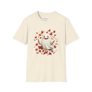 May include: Cream-colored t-shirt featuring a cartoon ghost with a smiling face, surrounded by autumn leaves in shades of red, orange, and brown. The ghost has outstretched arms. The shirt has a crew neck and short sleeves.