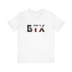 Może przedstawiać: Biała koszulka z nadrukiem liter "BTX" z przodu. Litery są w stylu vintage, z literą "B" z gwiazdą, a "T" i "X" z wzorem flagi.