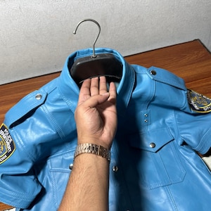 Peut inclure: Une chemise à manches courtes en cuir bleu clair avec un écusson de police sur la manche gauche. L'écusson porte les mots "Police Department" et "City of New York".