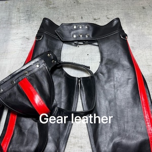 Puede incluir: Chaps de cuero negro con detalles rojos. Las chaps tienen cintura alta y se aseguran con una hebilla. El texto "Gear leather" es visible en la imagen.