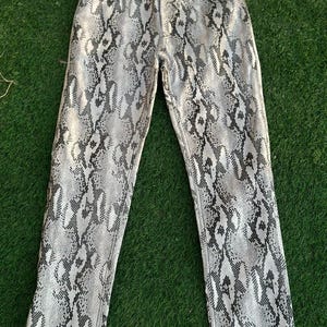 May include: A pair of straight-leg jeans with a snakeskin pattern in shades of gray and black. The jeans are laid flat on a green surface, showcasing the full length and details of the fabric.