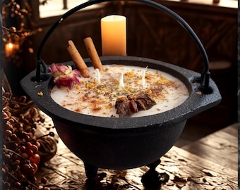 Puede incluir: Una vela en caldero de hierro fundido negro con tres mechas, decorada con ramas de canela, un anís estrellado y pétalos de flores secas. El texto 3-wick, Gauldron y CANDLE es visible. La vela se encuentra en un entorno oscuro con otras velas.