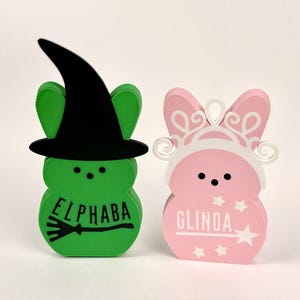 May include: Two green and pink bunny-shaped figures with painted faces and names. The green bunny is wearing a black witch hat and has the name "Elphaba" and a broom painted on it. The pink bunny is wearing a white crown and has the name "Glinda" and a star painted on it.