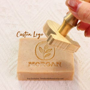 May include: A custom logo stamp is pressed onto a light brown bar of soap. The soap is stamped with a leaf design inside a circle and the name "MORGAN". The words "Custom Logo" are above the soap.