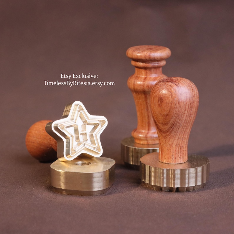 May include: Three wax seal stamps with wooden handles and brass bases. One stamp features a star-shaped design on its base. The text Etsy Exclusive: TimelessByRitesia.etsy.com is visible in the upper left area of the image. The stamps have polished metal bases and natural wood grain handles.