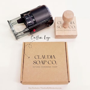Pode incluir: Um carimbo automático preto, um carimbo com cabo de madeira e uma caixa de papelão quadrada. O carimbo de madeira e a caixa apresentam o texto impresso em preto "CLAUDIA SOAP CO. NATURAL HANDMADE SOAP". Uma seta tracejada aponta para a caixa com "Custom Logo".
