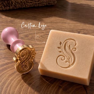 May include: A wax seal stamp with a pink handle and a gold-coloured brass head featuring an ornate letter S design. Next to it is a square block of soap with the same letter S embossed on its surface. The text Custom Logo is visible above.
