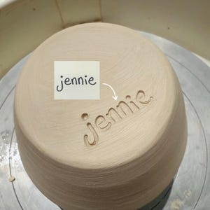 May include: A light beige, unglazed ceramic bowl with a smooth, textured surface. The name jennie is carved into the side of the bowl in a lowercase, cursive-style font. A small white square with the printed text jennie is placed above.