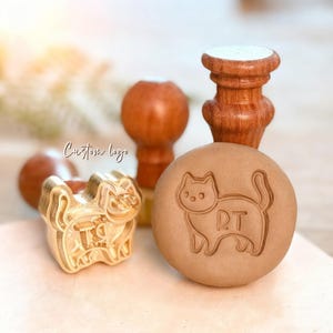 May include: A brass stamp head featuring a cat outline with the letters RT inside, positioned next to a round clay impression showing the same cat design. The stamp has a wooden handle, and the text Custom logo is visible in the background.