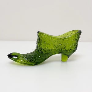 May include: A green glass decorative boot with a textured, starburst pattern. The boot has a pointed toe and a small heel. The glass is a deep, rich green color, and the boot is likely a decorative item.