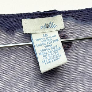 May include: A dark blue mesh garment with a white clothing tag featuring the brand name Odille. The tag displays size 10, 100% nylon, 100% cotton lining, 100% silk trim, Made in India, RN 56170, CA 32054, and the text See Reverse For Care.