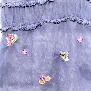 May include: A periwinkle blue mesh dress featuring horizontal ruffled tiers and scattered embroidery. The fabric is adorned with small, colorful floral motifs and detailed butterfly designs in shades of pink, purple, orange, and green.