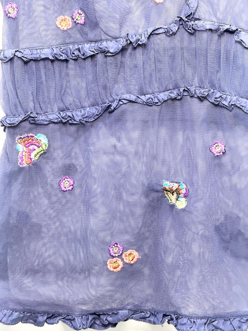 May include: A periwinkle blue mesh dress featuring horizontal ruffled tiers and scattered embroidery. The fabric is adorned with small, colorful floral motifs and detailed butterfly designs in shades of pink, purple, orange, and green.