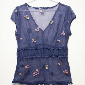 May include: A sheer navy blue short-sleeve top with a V-neckline, featuring small embroidered floral and butterfly motifs in orange, purple, and green. The garment includes a horizontal ruffled band across the midsection and ruffled trim edges.