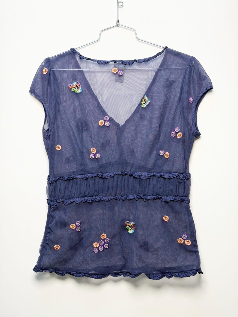 May include: A sheer navy blue short-sleeve top with a V-neckline, featuring small embroidered floral and butterfly motifs in orange, purple, and green. The garment includes a horizontal ruffled band across the midsection and ruffled trim edges.