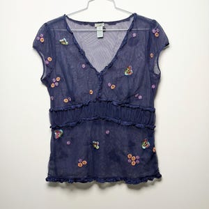 May include: A dark blue sheer mesh top with short sleeves and a V-neckline. The garment features small embroidered floral patterns and colorful butterfly motifs scattered across the front, with ruffled trim along the neckline and waistline.