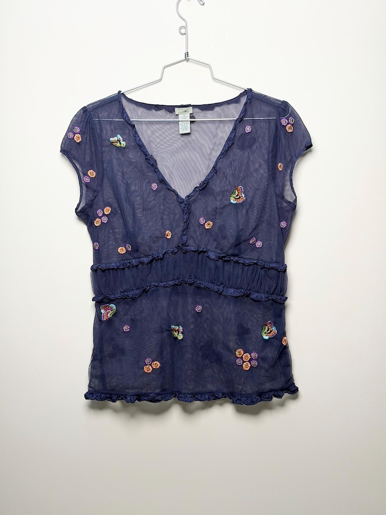 May include: A dark blue sheer mesh top with short sleeves and a V-neckline. The garment features small embroidered floral patterns and colorful butterfly motifs scattered across the front, with ruffled trim along the neckline and waistline.