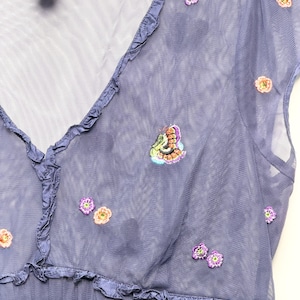 May include: A periwinkle blue mesh top featuring a V-neckline with ruffled trim and short sleeves. The sheer fabric is decorated with small, scattered embroidered floral motifs in shades of purple, orange, and green, along with a butterfly detail.