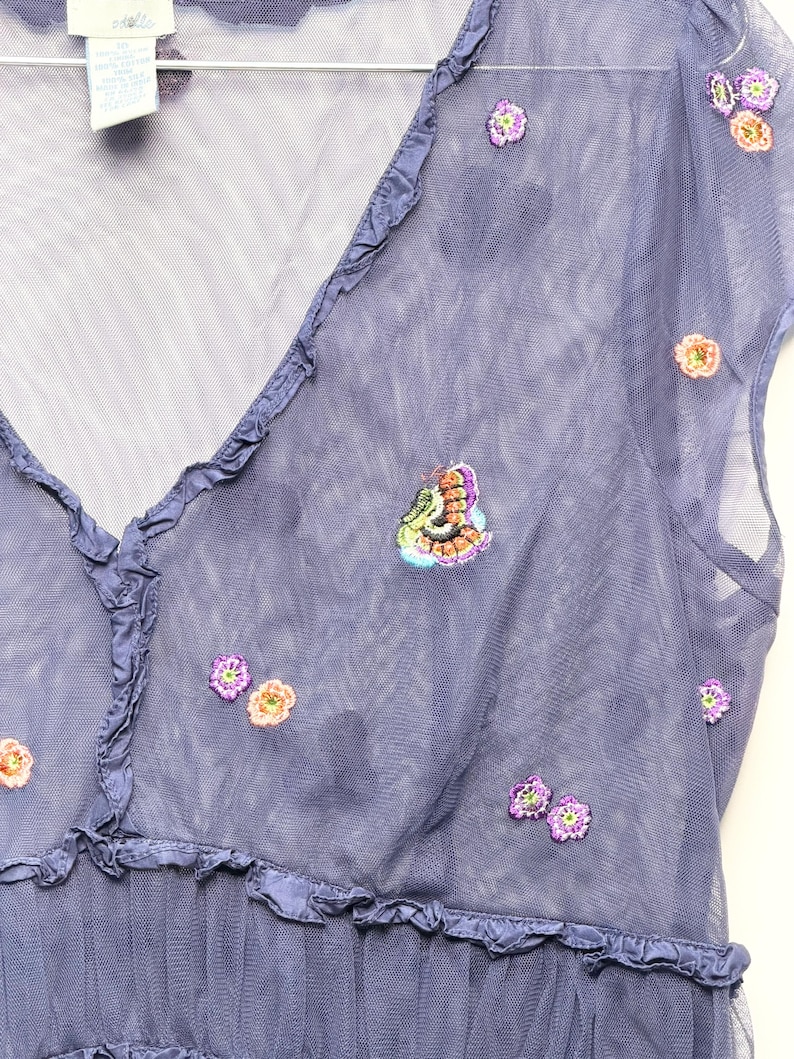 May include: A periwinkle blue mesh top featuring a V-neckline with ruffled trim and short sleeves. The sheer fabric is decorated with small, scattered embroidered floral motifs in shades of purple, orange, and green, along with a butterfly detail.