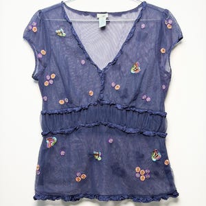 May include: A sheer navy blue V-neck top with short sleeves, featuring a ruffled empire waist and hem. The garment is decorated with small, colorful embroidered butterfly and flower motifs scattered across the front and sleeves.