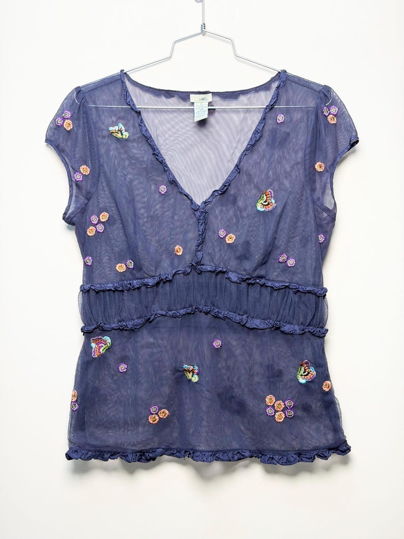 May include: A sheer navy blue V-neck top with short sleeves, featuring a ruffled empire waist and hem. The garment is decorated with small, colorful embroidered butterfly and flower motifs scattered across the front and sleeves.