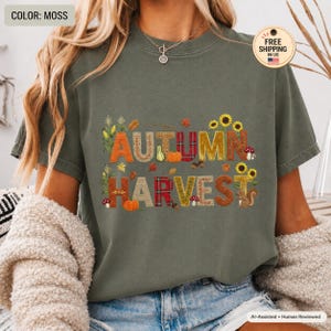 Puede incluir: Una camiseta de manga corta color verde musgo con el texto AUTUMN HARVEST en letras grandes. Las letras están decoradas con motivos de calabazas, girasoles, setas y una ardilla sobre un fondo de color verde oscuro sólido.