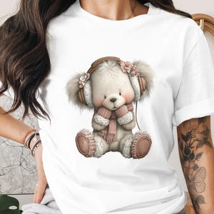 Könnte beinhalten: Weißes T-Shirt mit einem Cartoon-Bären-Design. Der Bär trägt Ohrenschützer mit Blumen, einen rosa Schal und Kopfhörer. Der Bär sitzt mit verschränkten Beinen. Das Design verwendet weiche Pastellfarben.