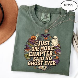 Op de afbeelding: Een mosgroen t-shirt met een grafische print met de tekst "JUST ONE MORE CHAPTER... SAID NO GHOST EVER". Het ontwerp bevat een geest met een bril, stapels boeken, een pompoen, een heksenhoed, een vleermuis, een ketel en spinnenwebben.