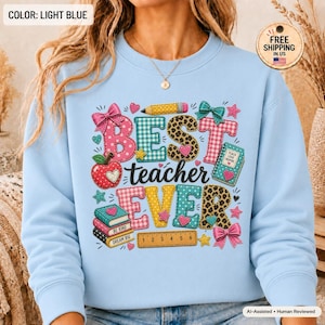 Peut inclure: Un sweat-shirt bleu clair avec le texte BEST teacher EVER dans des motifs colorés. Le design comprend un crayon, une pomme rouge, des livres empilés, une règle et des nœuds. Le texte est décoré de motifs léopard, carreaux et pois.