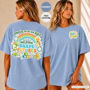 Puede incluir: Una camiseta azul claro con un gráfico colorido en la espalda que muestra un arcoíris, bloques de letras, un portapapeles, una bombilla y un silbato. El texto dice PROUD TO BE AN RBT HELPING SHAPE FUTURES. El frente tiene un logo RBT TEAM.